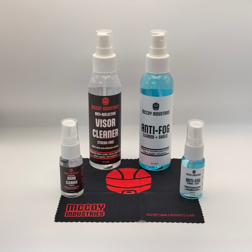 A set of McCoy Industries visor cleaning products arranged on a black microfiber cloth. The set includes a large bottle of Visor Cleaner, a large bottle of Anti-Fog Cleaner + Shield, a small bottle of Lens Cleaner, and a small bottle of Anti-Fog, with a red helmet graphic printed on the cloth.